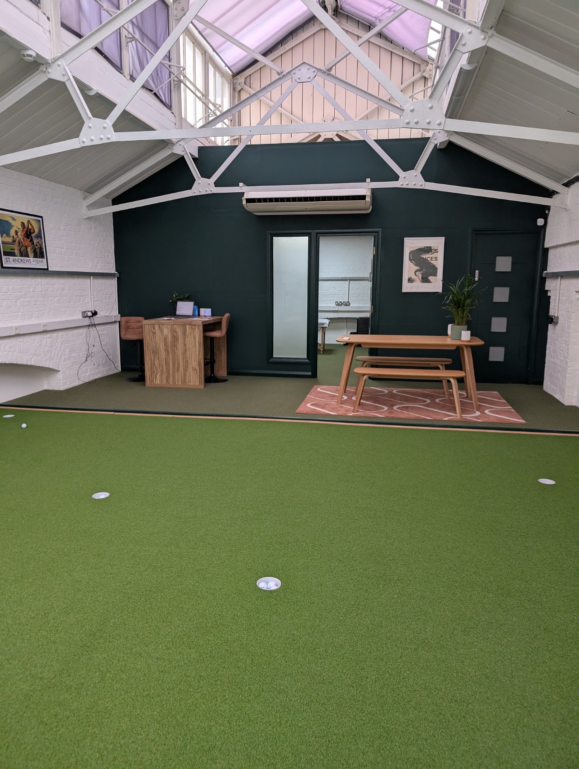 Indoor putting green and seating area beneath exposed white roof beams at The City Golf Club in Woolwich